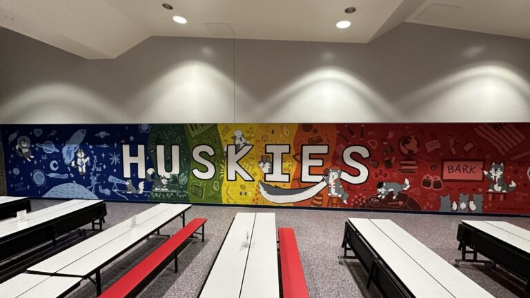 Zoomed out photo of a wall mural that is rainbow and says Huskies. There are paintings of huskies on it doing various jobs.